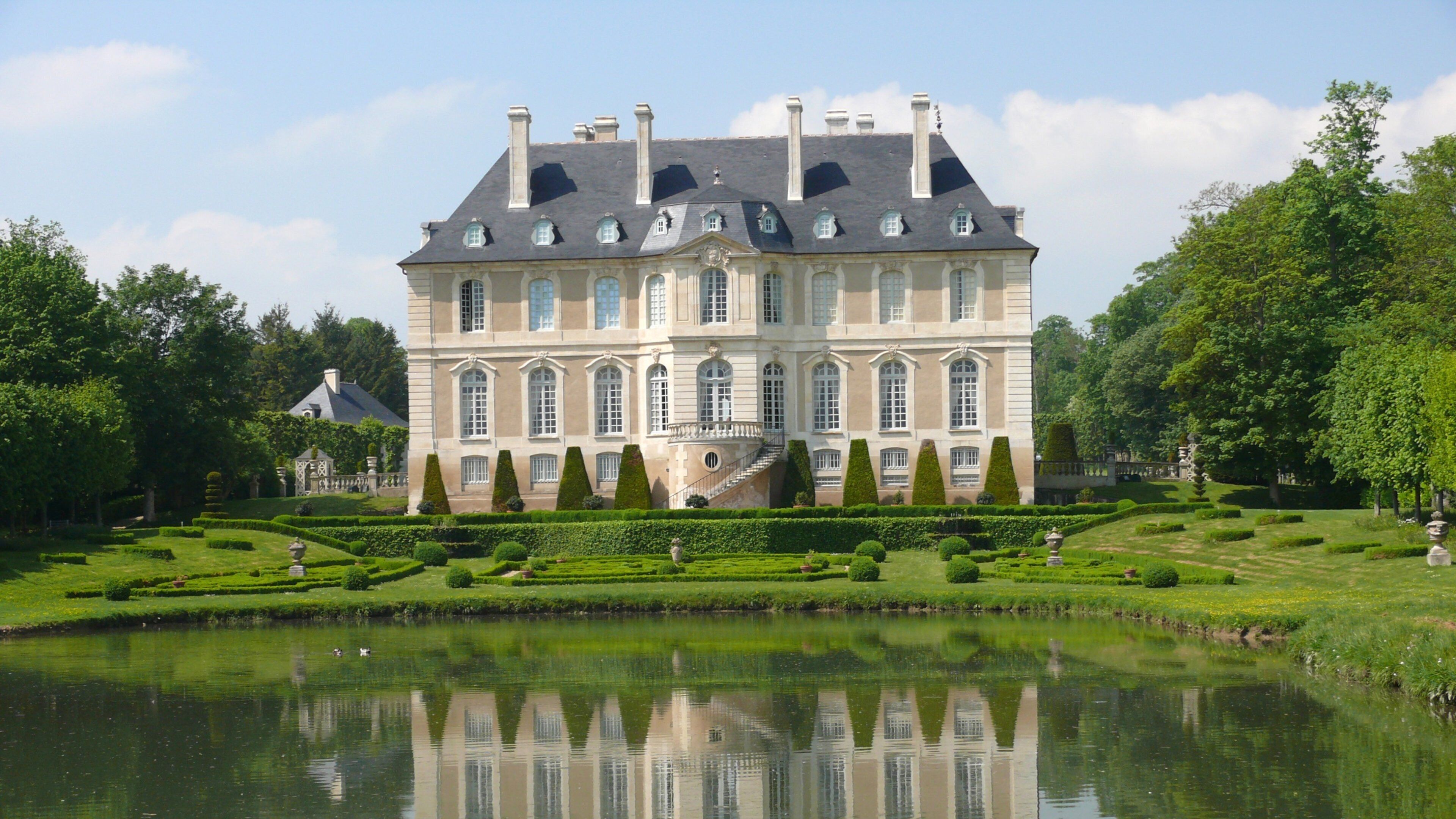 A large, historic building with a symmetrical facade, a central staircase, and a surrounding formal garden with a pond.