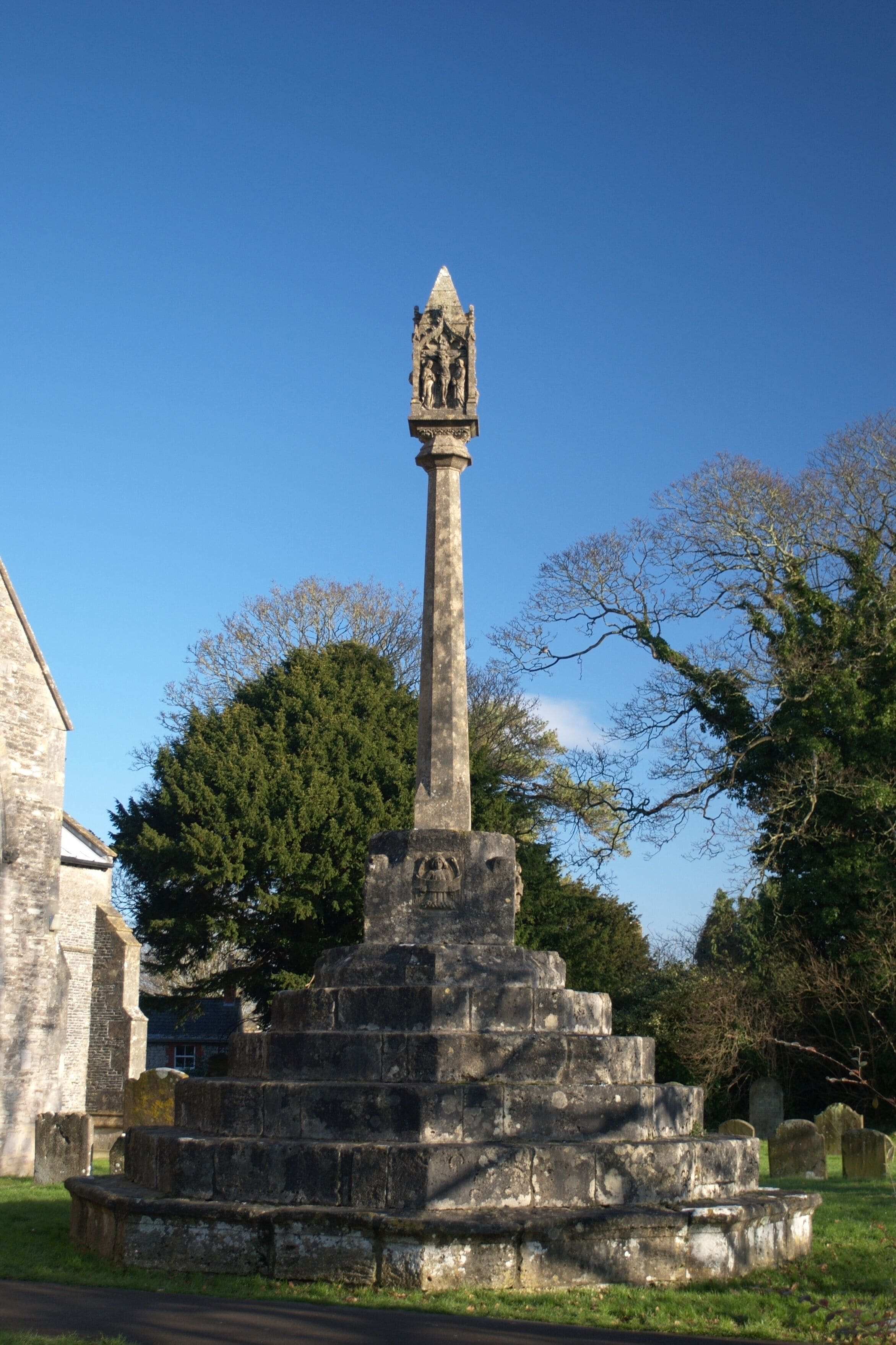 A stone monument with a statue on top, surrounded by trees and a building in the background.