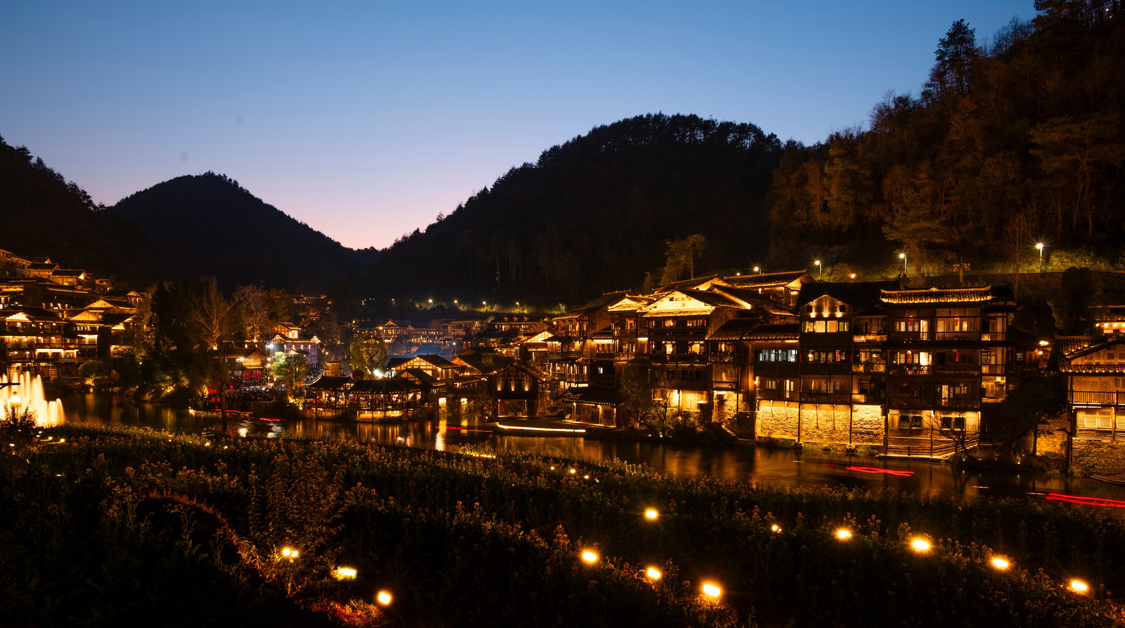 A traditional village at dusk with illuminated buildings and a calm river.
