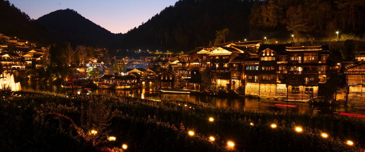A traditional village at dusk with illuminated buildings and a calm river.