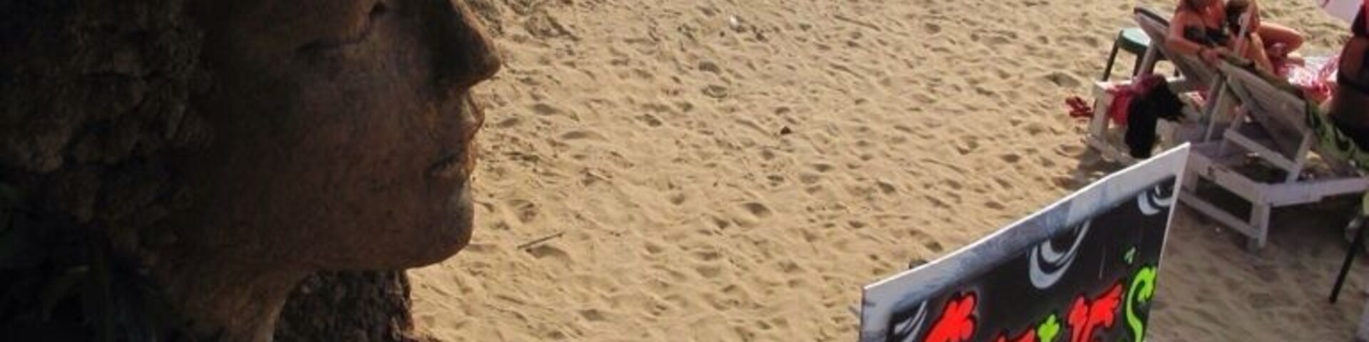 Una playa con gente relajándose bajo sombrillas, un cartel de 'Curry's' y barcos al fondo.