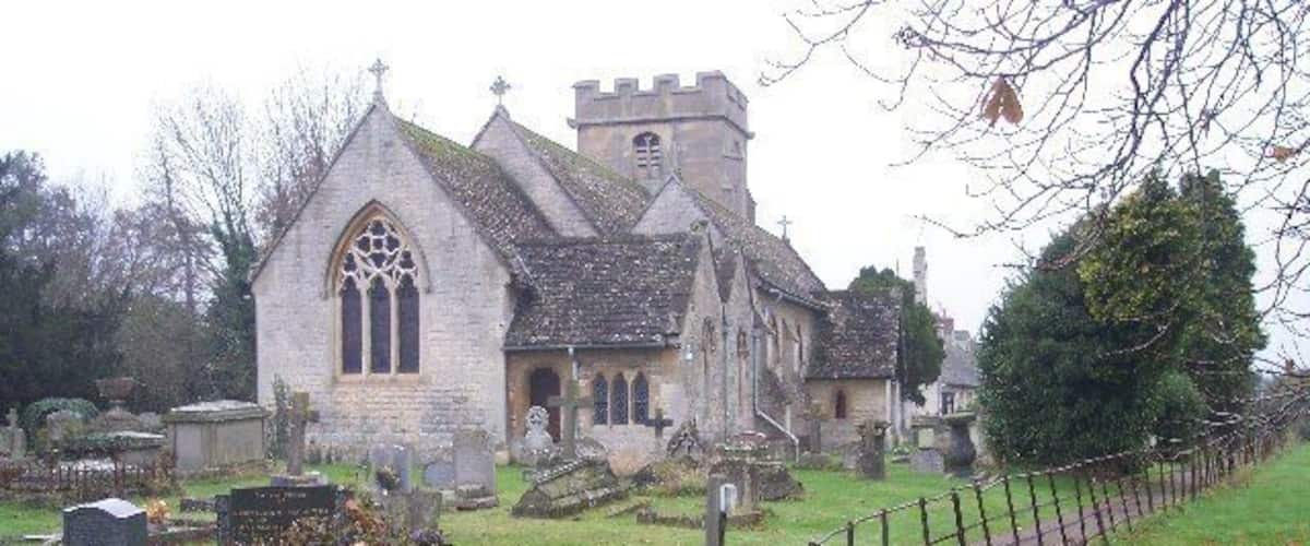 A church with a stone facade and a tall steeple, surrounded by a cemetery with headstones and a wooden fence.