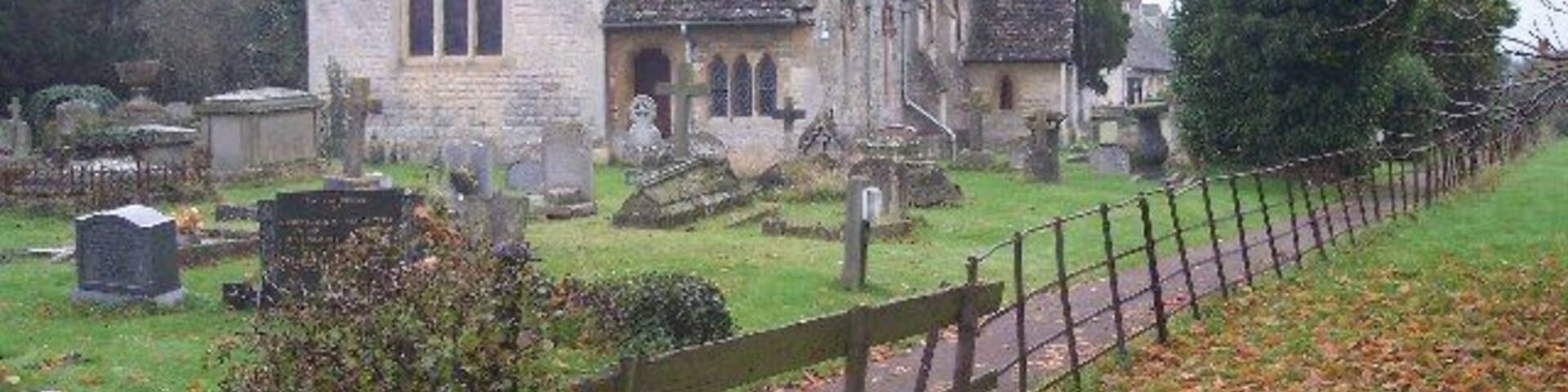 A church with a stone facade and a tall steeple, surrounded by a cemetery with headstones and a wooden fence.