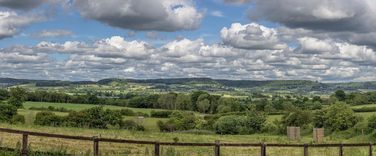 A rural landscape with rolling hills, a wooden fence, and a cloudy sky.