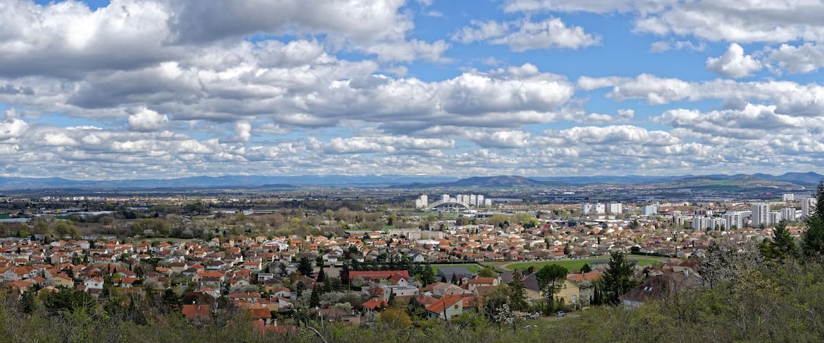 A cityscape with red-roofed houses, a distant mountain range, and a large body of water.
