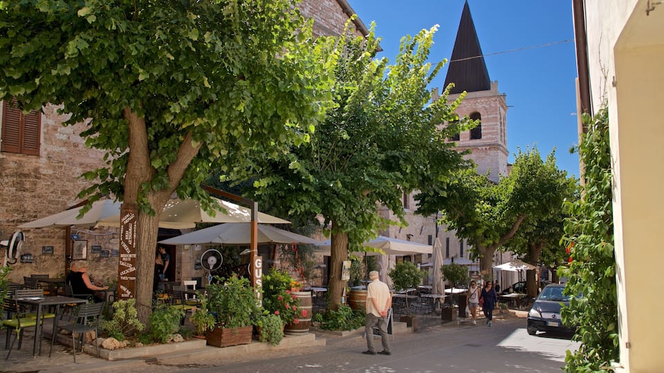 A narrow street lined with outdoor cafes, trees, and a church with a tall spire.