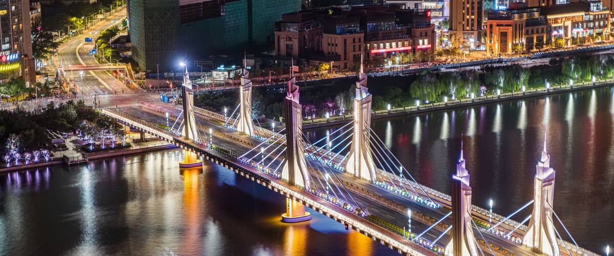 A brightly lit bridge with multiple towers over a calm river, surrounded by city buildings and a park with illuminated trees.
