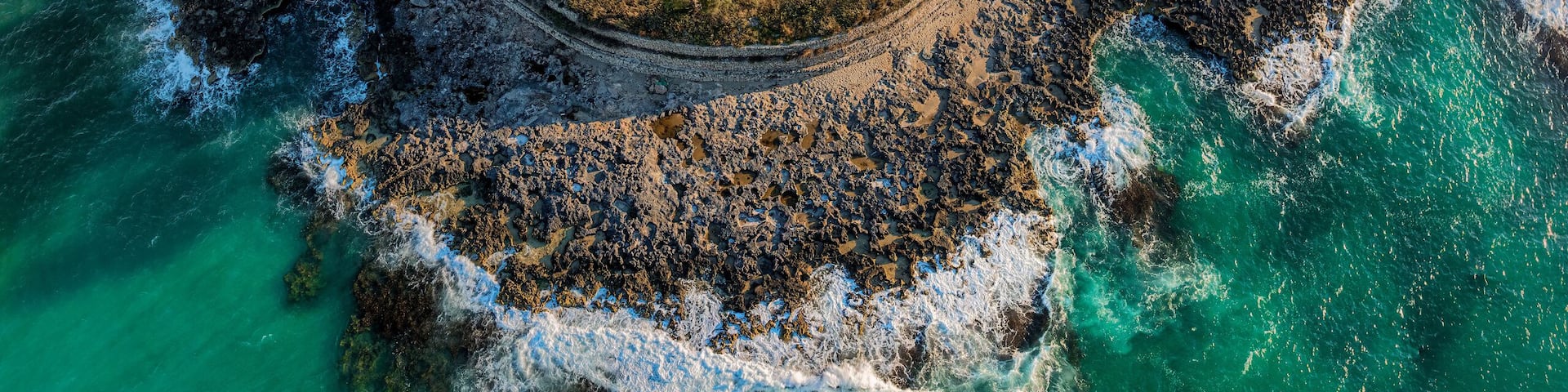 An aerial view of a coastal archaeological site with ruins, a surrounding grassy area, and a rocky coastline.