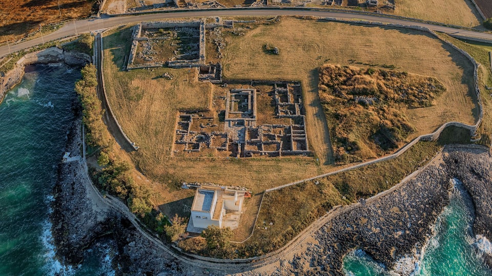 An aerial view of a coastal archaeological site with ruins, a surrounding grassy area, and a rocky coastline.