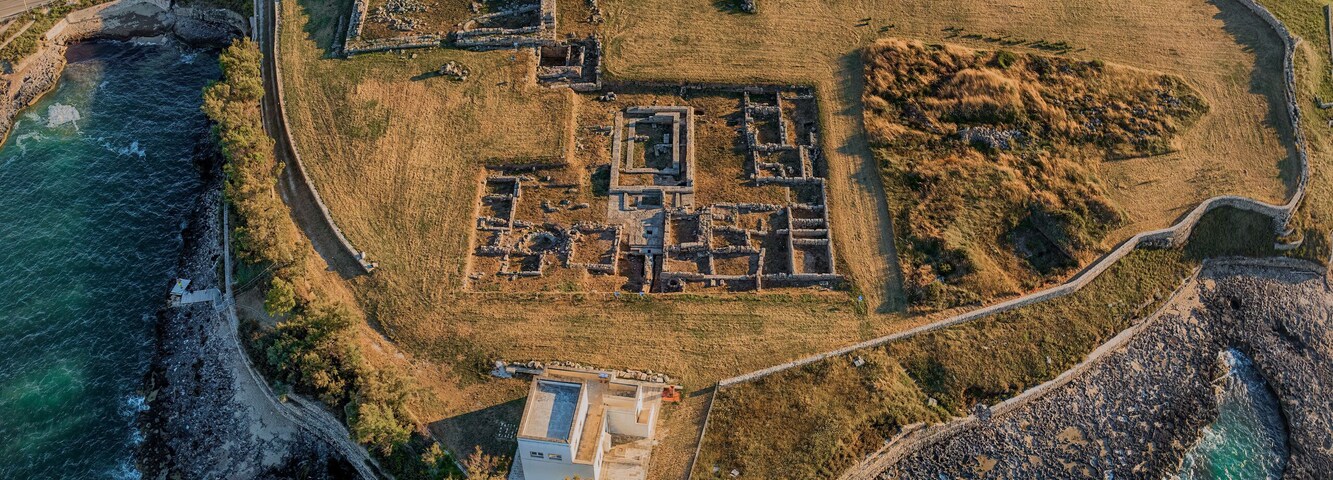 Vista aérea de un sitio arqueológico costero con ruinas, una zona verde circundante y un litoral rocoso.