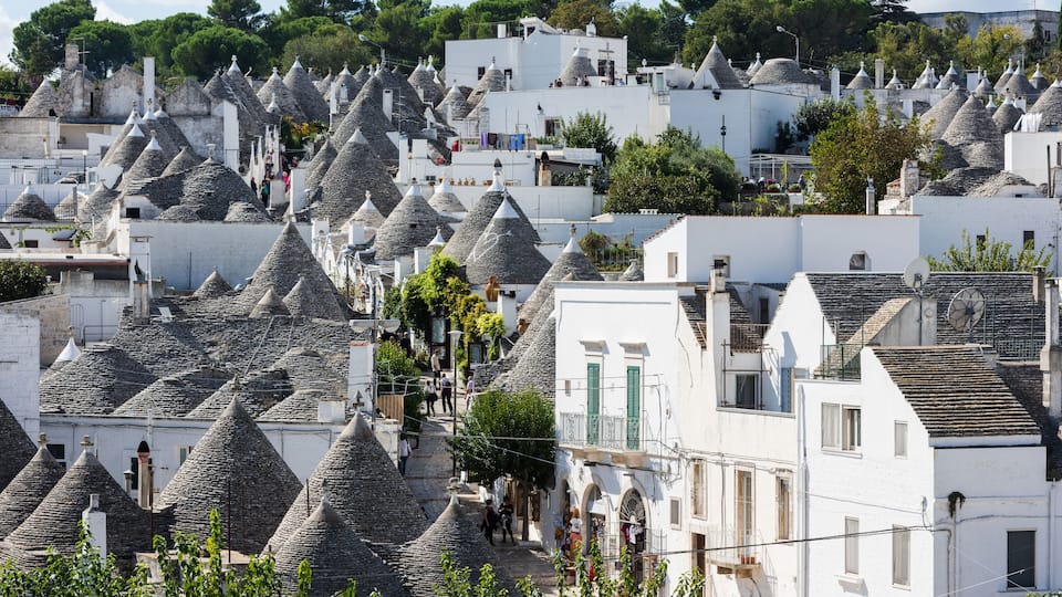 A village with white buildings and conical roofs, surrounded by trees and a hilly landscape.