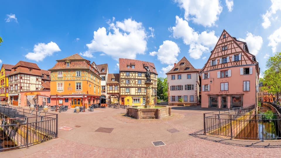 A historic town square with traditional half-timbered buildings, a fountain, and a canal.