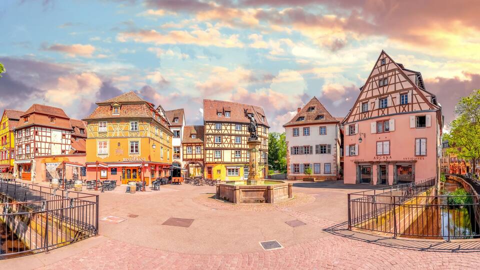 A European town square with traditional half-timbered buildings, a central fountain, and a canal.