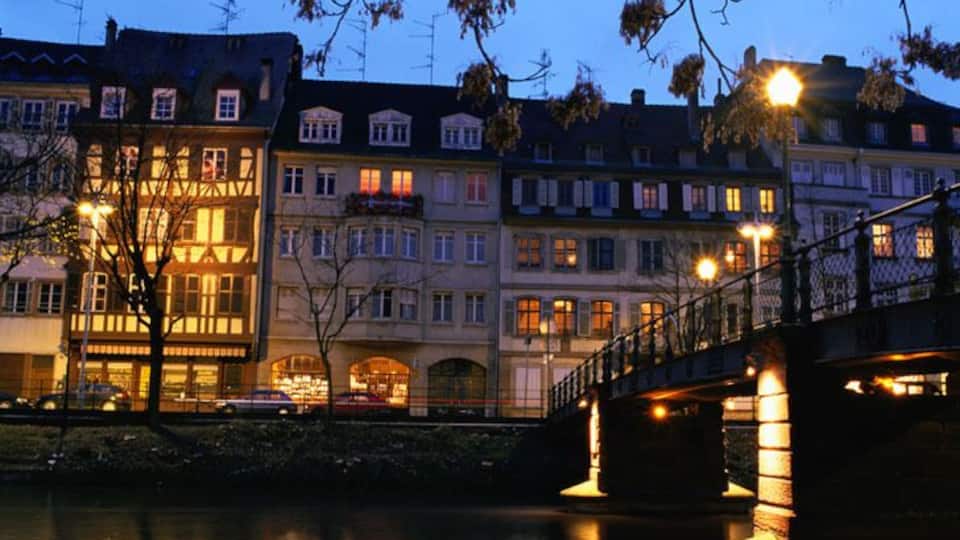 A riverside view of a European city at dusk with illuminated buildings and a bridge.