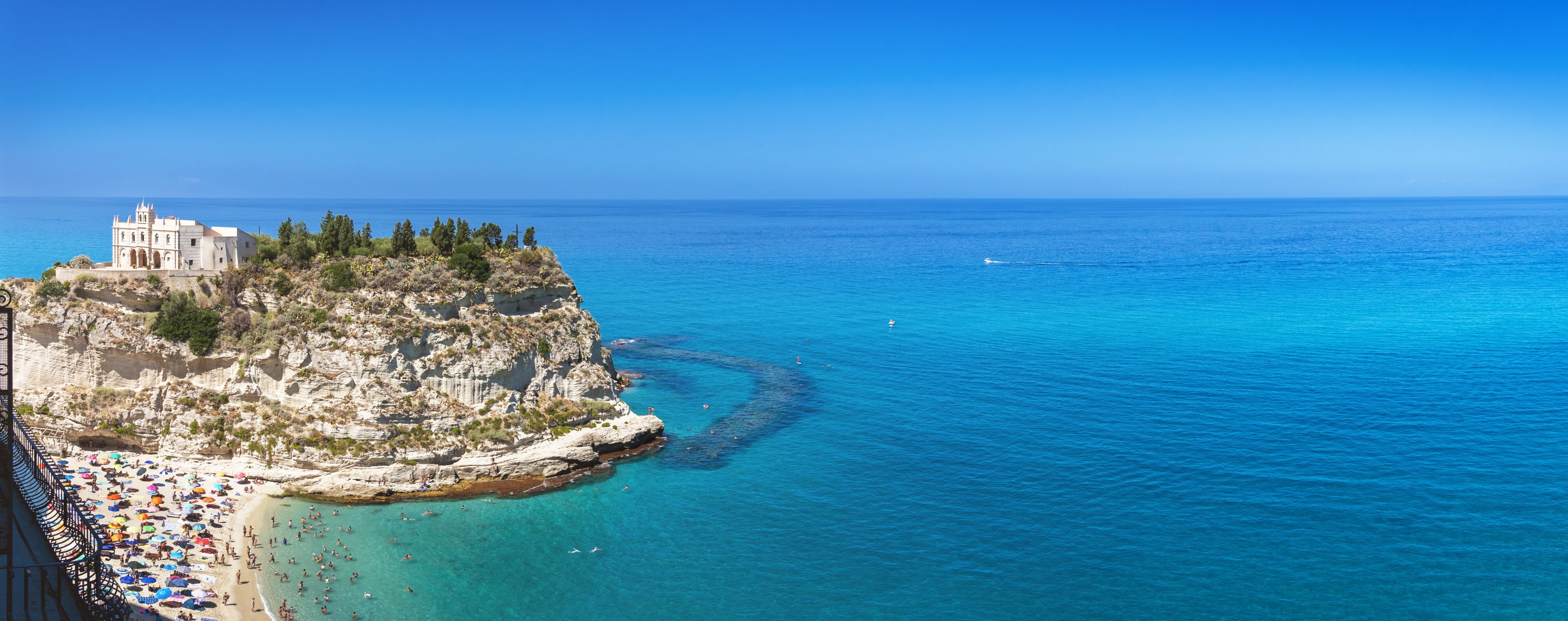 Uma cidade costeira com uma igreja histórica em um penhasco, uma praia com guarda-sóis e águas azul-claras.