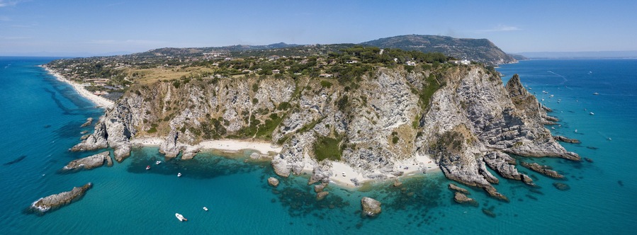 Un paisaje costero con una península rocosa, aguas turquesas cristalinas y un pueblo lejano.