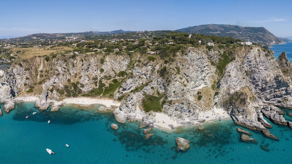 A coastal landscape with a rocky peninsula, clear turquoise waters, and a distant town.