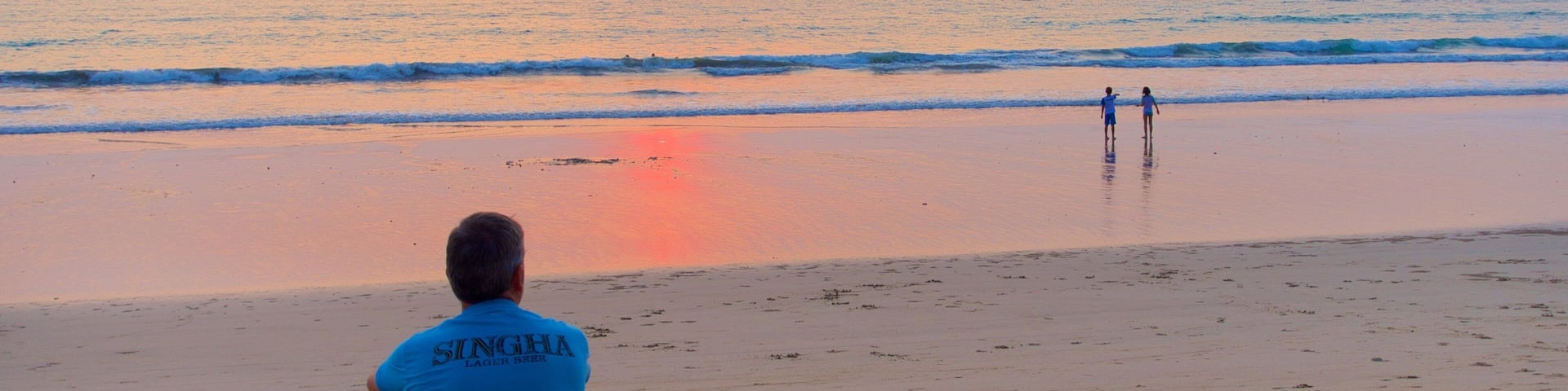 A beach at sunset with a person sitting and two people walking in the distance.
