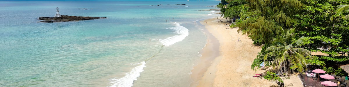 Une plage aux eaux turquoise et limpides, un rivage sablonneux et un phare qui se dessine à l’horizon.