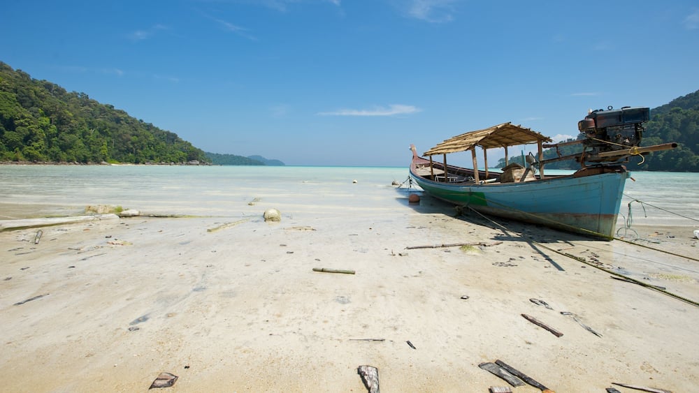 A boat on a sandy beach with clear turquoise water and a forested hill in the background.