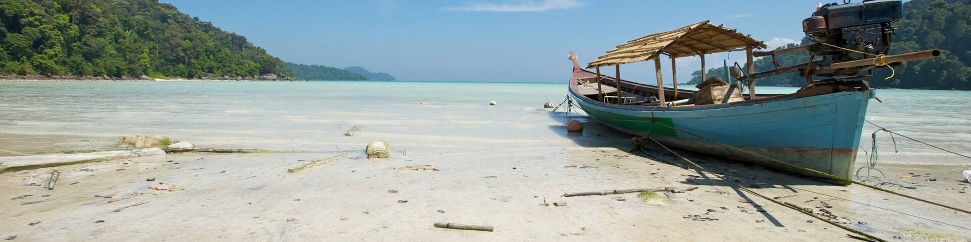 A boat on a sandy beach with clear turquoise water and a forested hill in the background.
