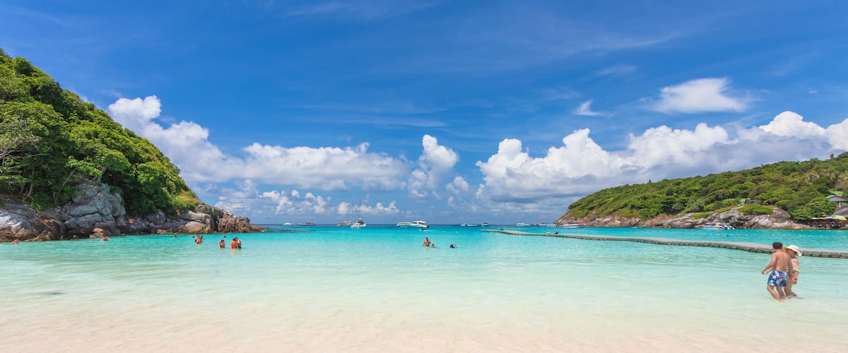 A beach with clear turquoise water, people swimming, and boats in the distance.