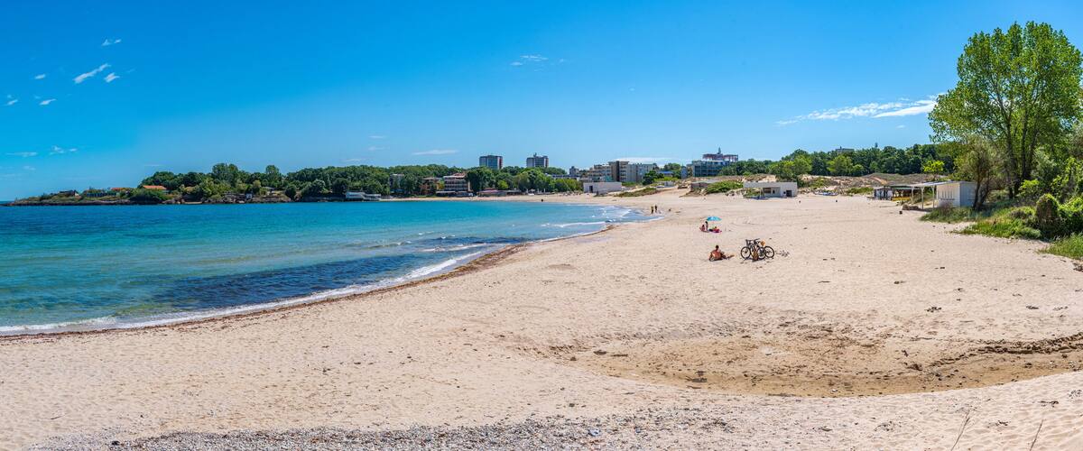 A beach with clear blue water, a sandy shore, and buildings in the background.
