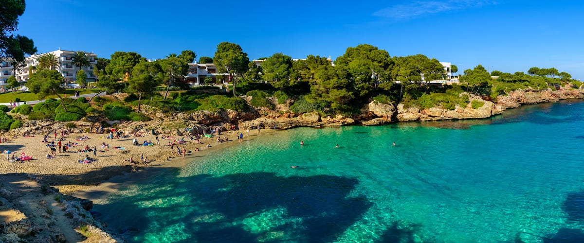 A coastal beach with clear turquoise waters, a sandy shore, and a backdrop of lush greenery and buildings.