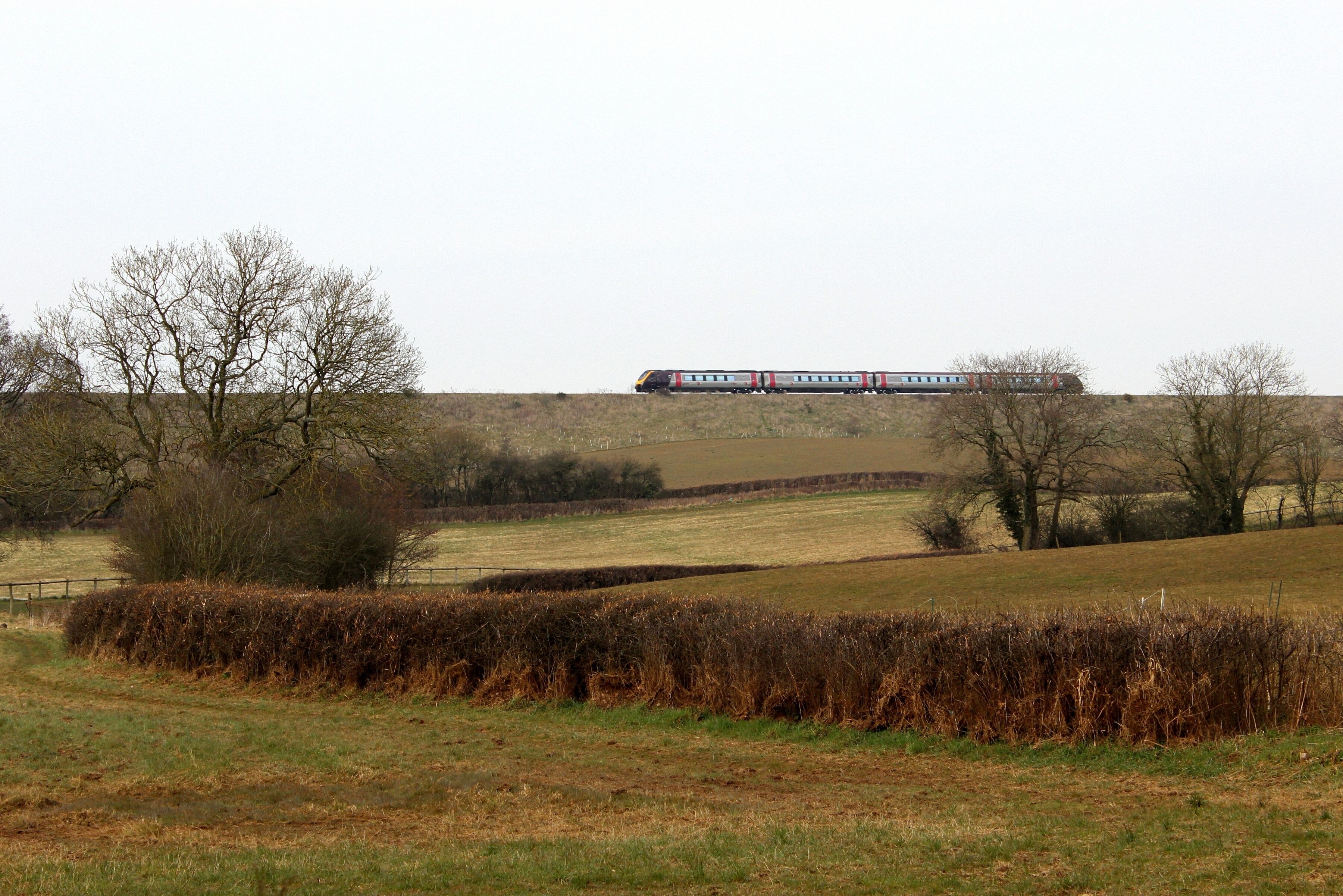 A train on a track in a rural landscape with fields, trees, and a fence.