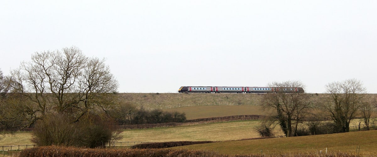 A train on a track in a rural landscape with fields, trees, and a fence.