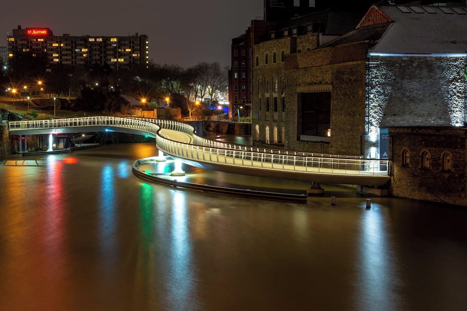 Een gebogen brug over het water, 's nachts verlicht met reflecties op het water. Achtergrond toont gebouwen en een Marriott hotel.