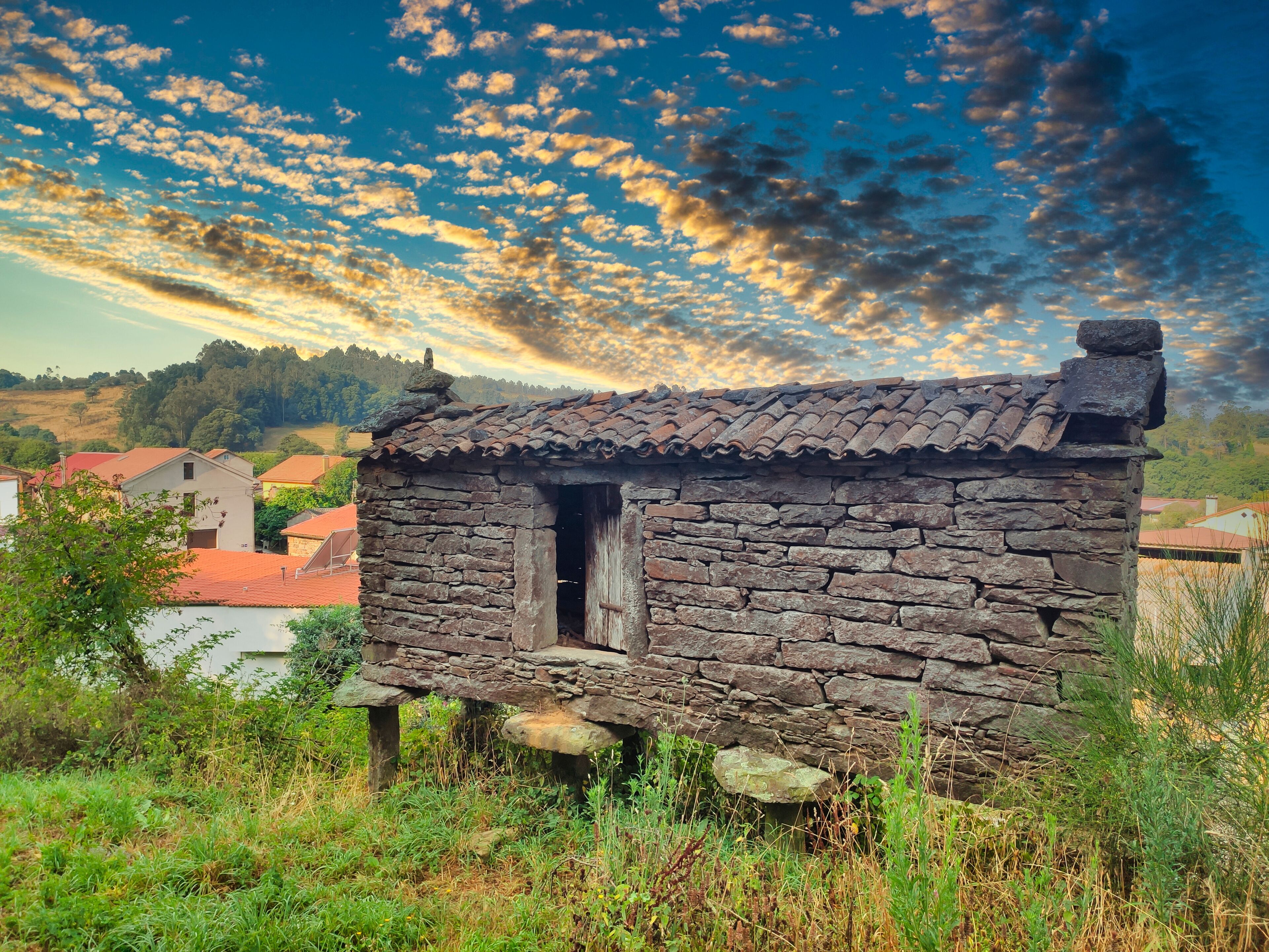 A rustic stone cottage with a tiled roof, surrounded by greenery and set against a backdrop of a clear sky with scattered clouds.