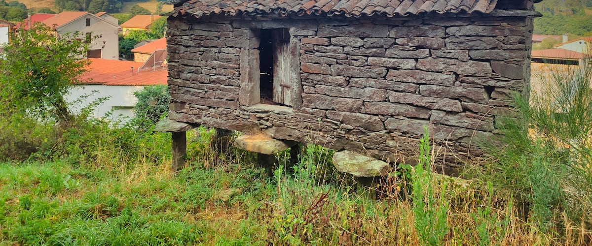 A rustic stone cottage with a tiled roof, surrounded by greenery and set against a backdrop of a clear sky with scattered clouds.