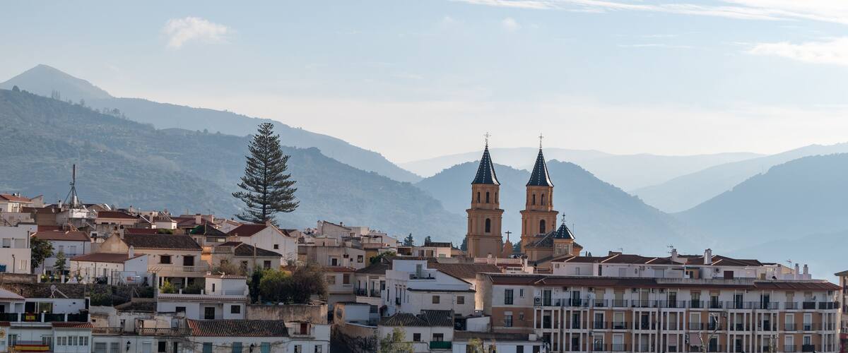 A town with two prominent church towers, surrounded by residential buildings and mountains in the background.