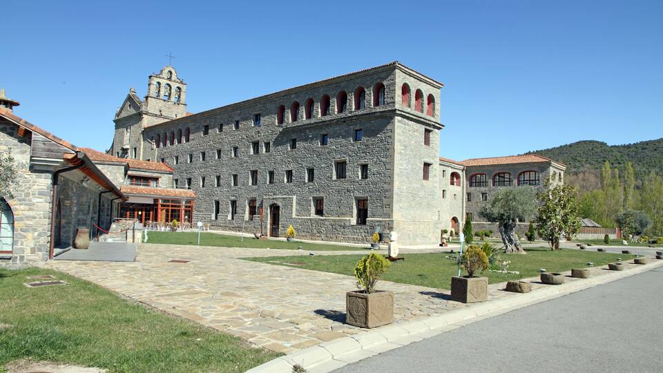 A historic stone building with a bell tower, a paved courtyard, and a hillside in the background.