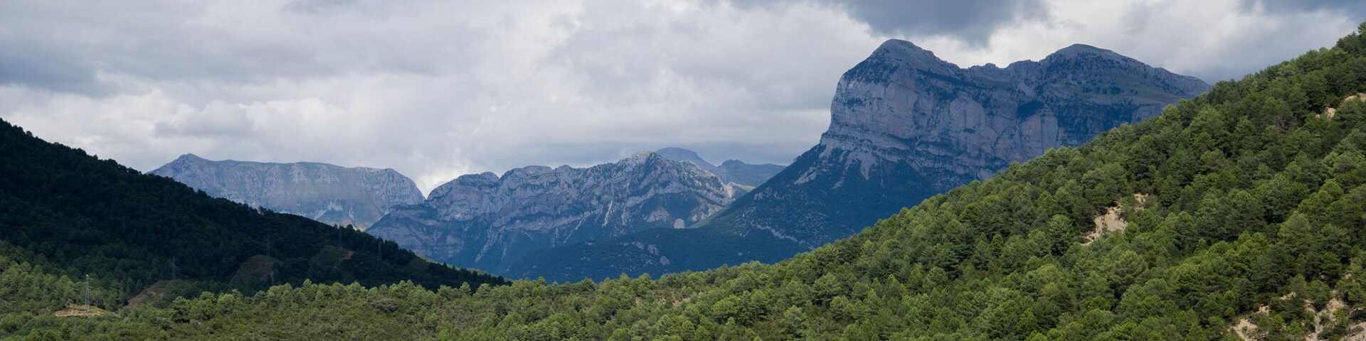 A mountainous landscape with dense green forests and rugged peaks under a cloudy sky.