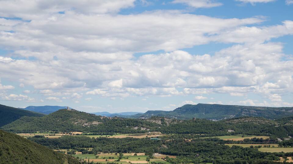 A mountainous landscape with rolling hills, scattered trees, and a clear sky with clouds.