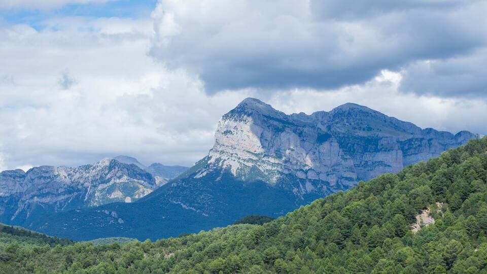 A mountain range with a prominent peak, dense forested hills, and a cloudy sky.