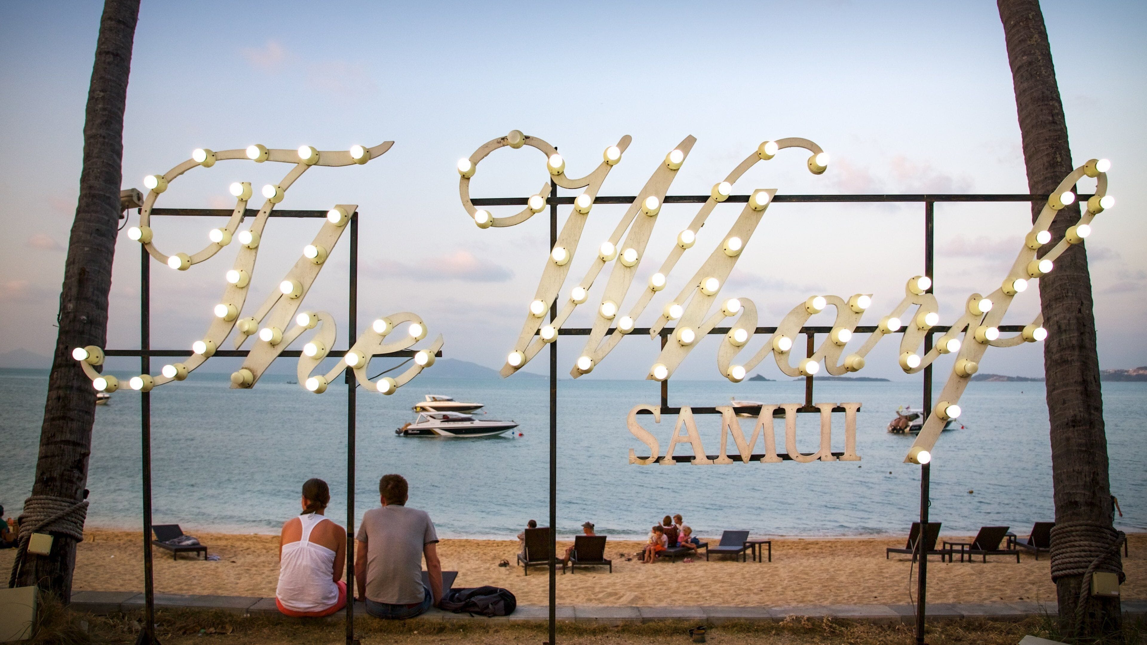 A beach with palm trees, a boat on the water, and a sign with lights spelling out "The Big Samu".