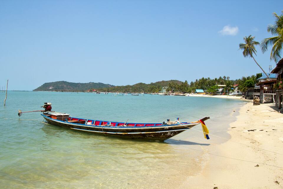 A boat on a beach with clear water, palm trees, and a distant hill.