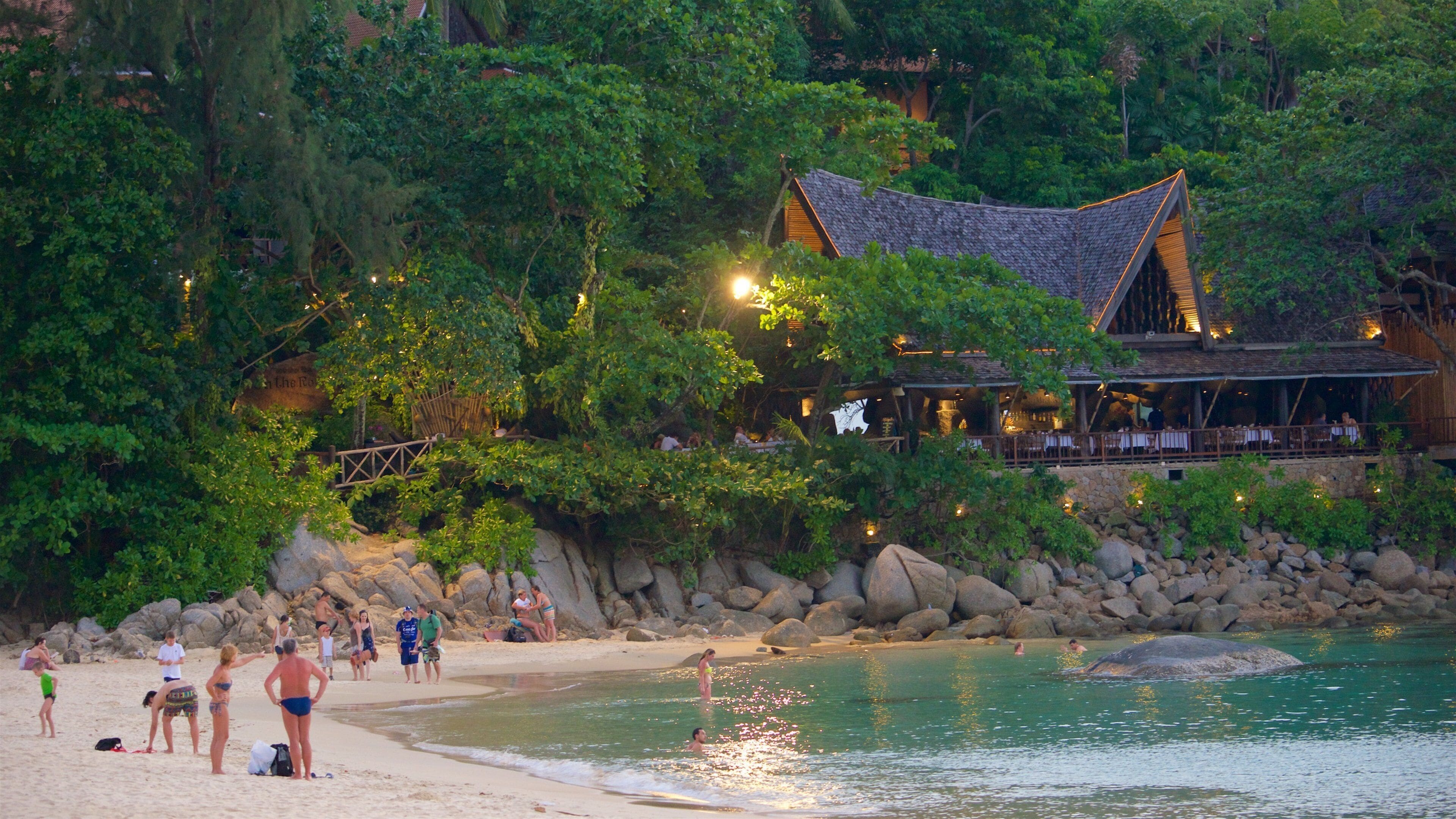 A beach with people, a restaurant with a traditional roof, and a rocky shoreline.