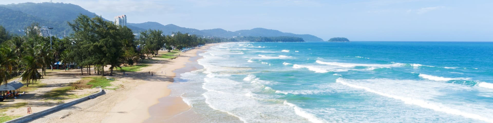 A beach with clear blue water, sandy shore, palm trees, and mountains in the background.