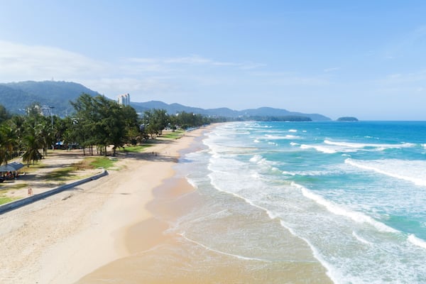 Ein Strand mit klarem, blauem Wasser, sandigem Ufer, Palmen und Bergen im Hintergrund.