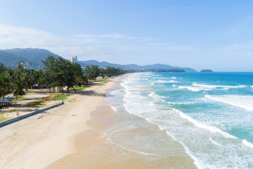 Een strand met helderblauw water, een zandstrand, palmbomen en bergen op de achtergrond.