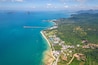 An aerial view of a coastal area with a clear blue sea, a long pier, and a densely forested landscape with buildings and a resort.