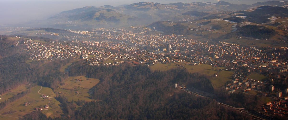 A panoramic view of a town nestled in a valley with surrounding mountains and a distant snow-capped range.