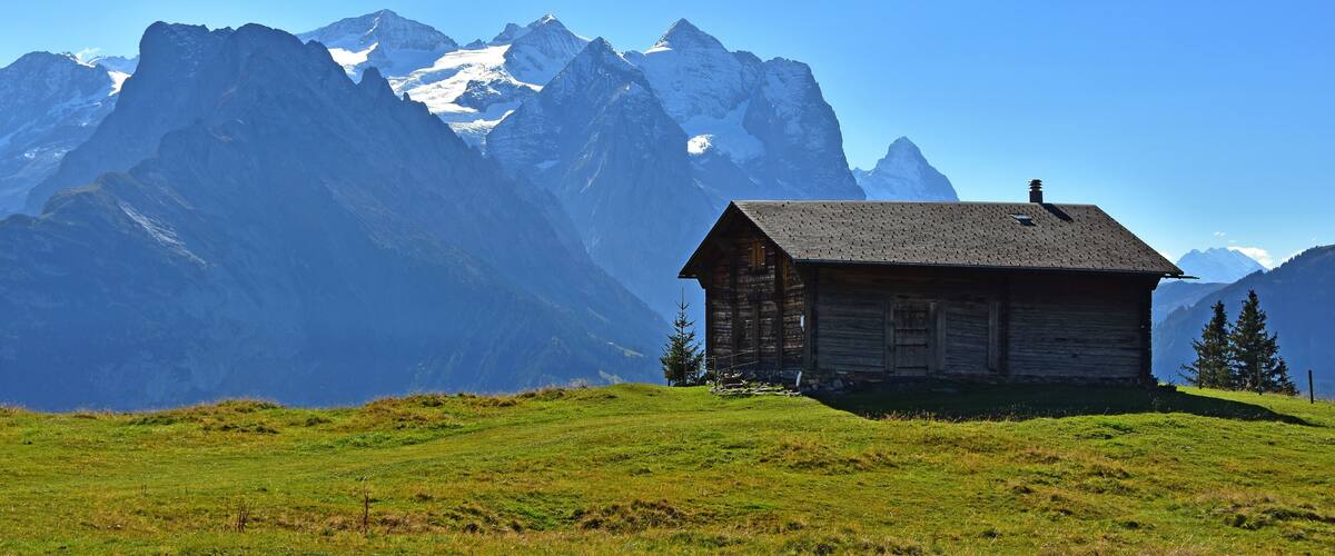 A wooden cabin on a grassy hill with snow-capped mountains in the background.