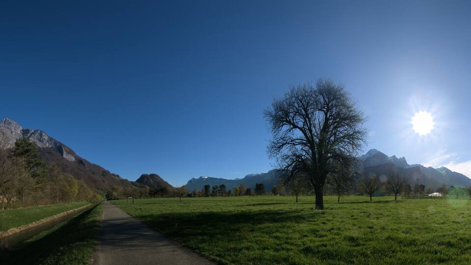 A rural landscape with a clear path, green fields, a river, and mountains in the background.