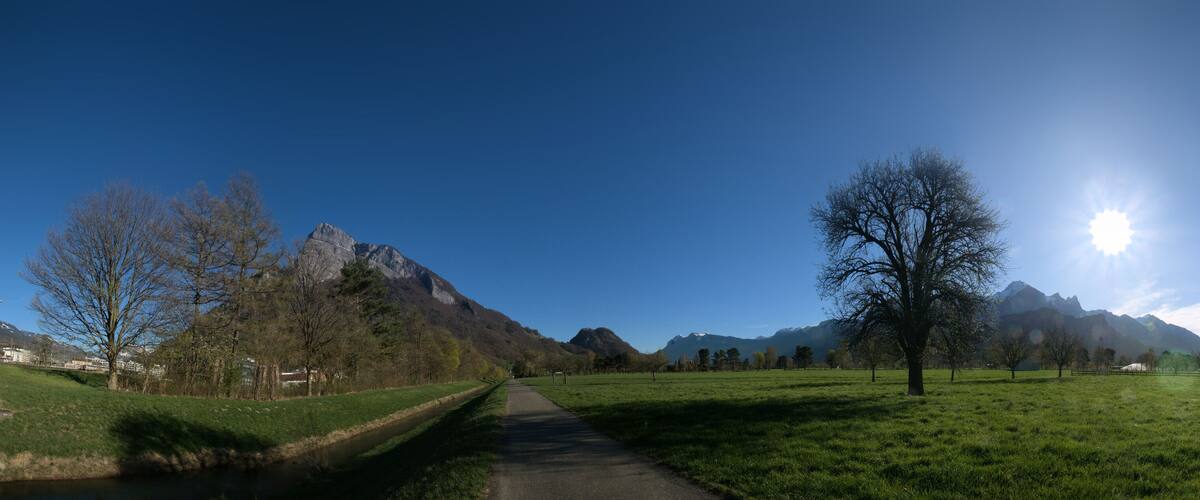 A rural landscape with a clear path, green fields, a river, and mountains in the background.