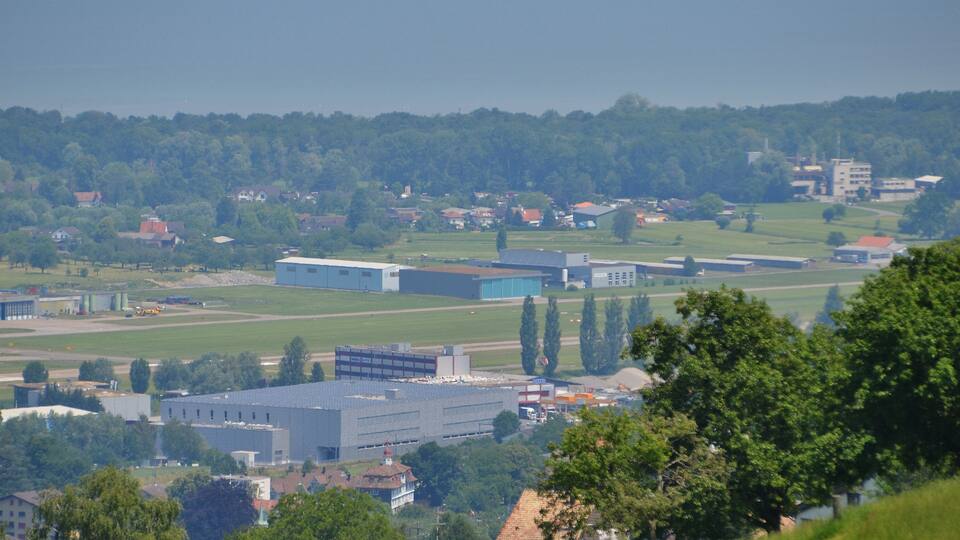 A landscape with industrial buildings, a large blue structure, and a forested hill in the background.
